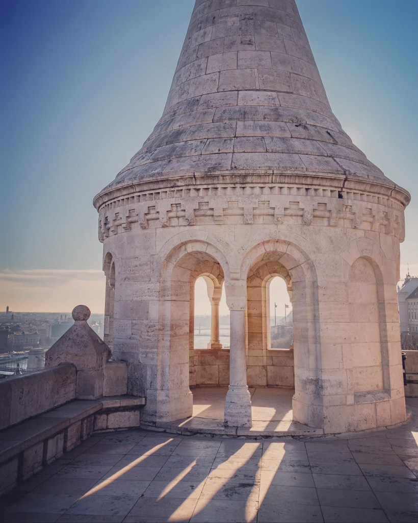 Close-up of a turret lookout at Fisherman’s Bastion with light streaming through arched windows and a distant city view.