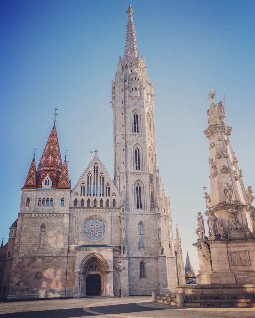 Exterior of Matthias Church with its towering spire, Gothic façade, and Baroque Trinity Column in front.
