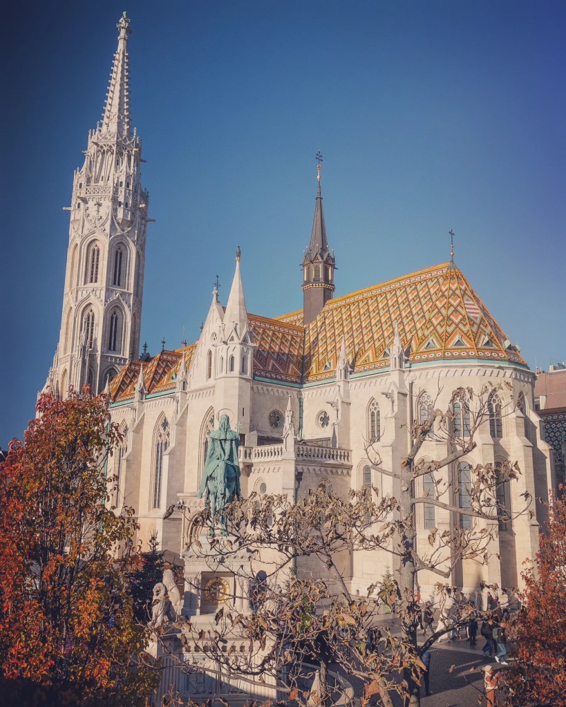 Side view of Matthias Church with its colorful Zsolnay tile roof, tall spire, and surrounding statue garden.