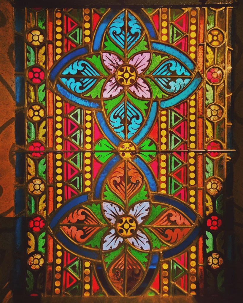 Vibrant stained glass window with geometric floral and leaf patterns in bright reds, blues, greens, and yellows.
