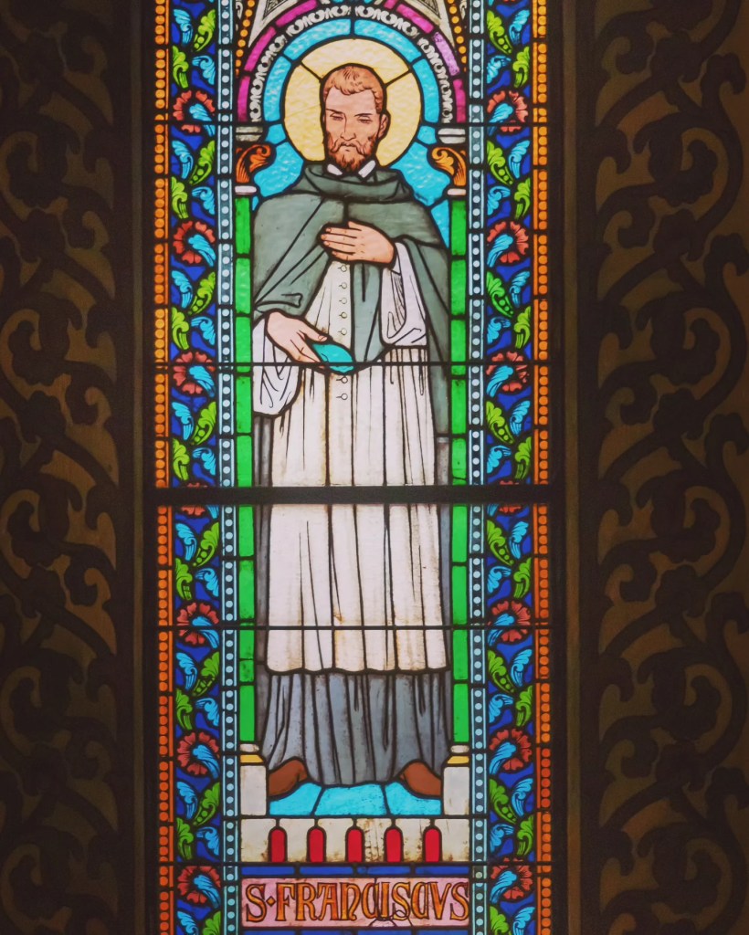 Stained glass window featuring Saint Franciscus, surrounded by colorful floral and border designs.