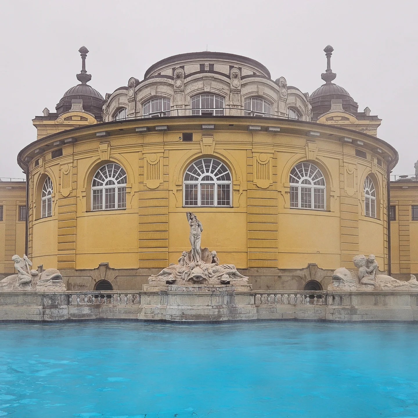 Neo-Baroque yellow bathhouse façade with statues and bright blue thermal water in front at Széchenyi Thermal Bath, Budapest