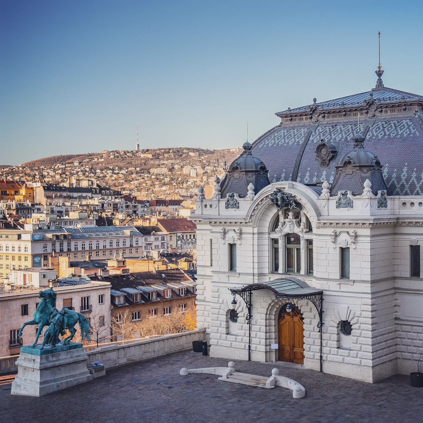 Historic white building with a decorative roof and horse statue on the Buda side of Budapest.