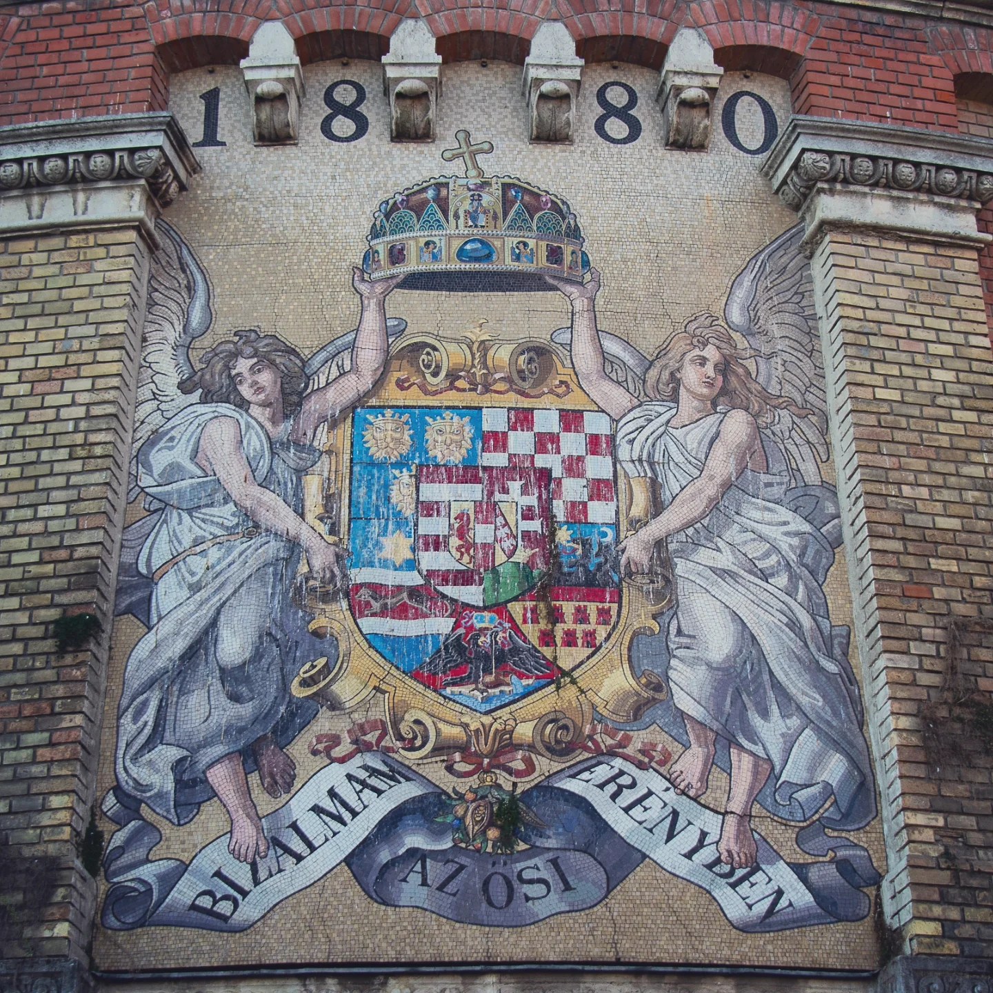Mosaic of Hungary’s historic coat of arms flanked by angels on a brick wall, dated 1880.
