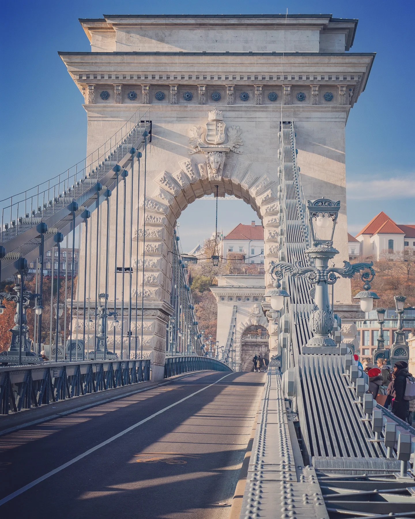 A view down the pedestrian walkway of the Széchenyi Chain Bridge with ornate ironwork and the Buda Castle Hill in the distance.