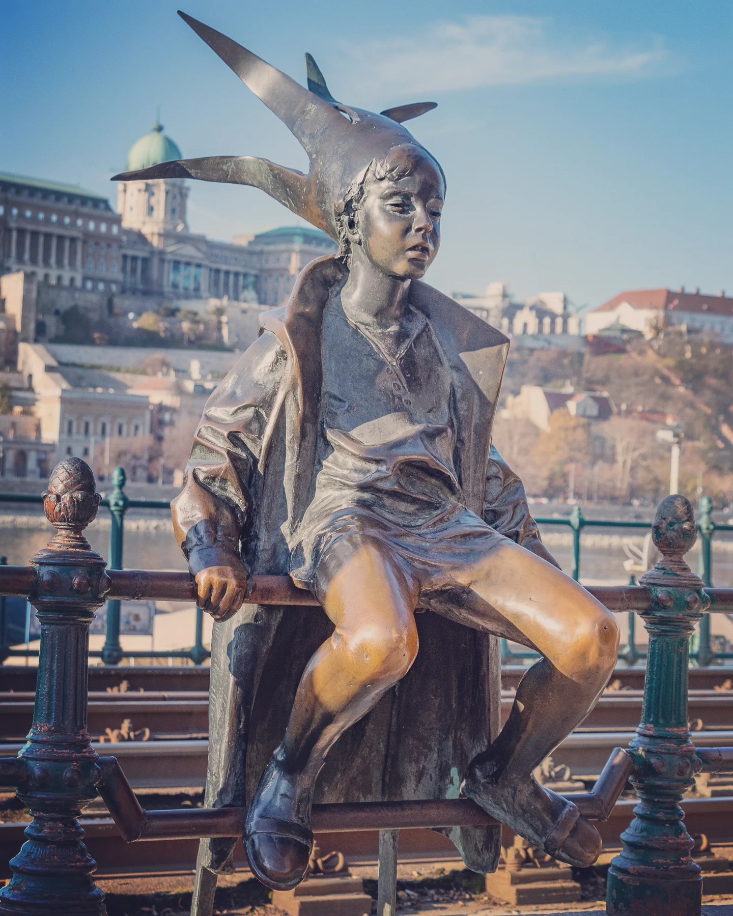 Bronze statue of a child wearing a crown, sitting on a railing beside the Danube.