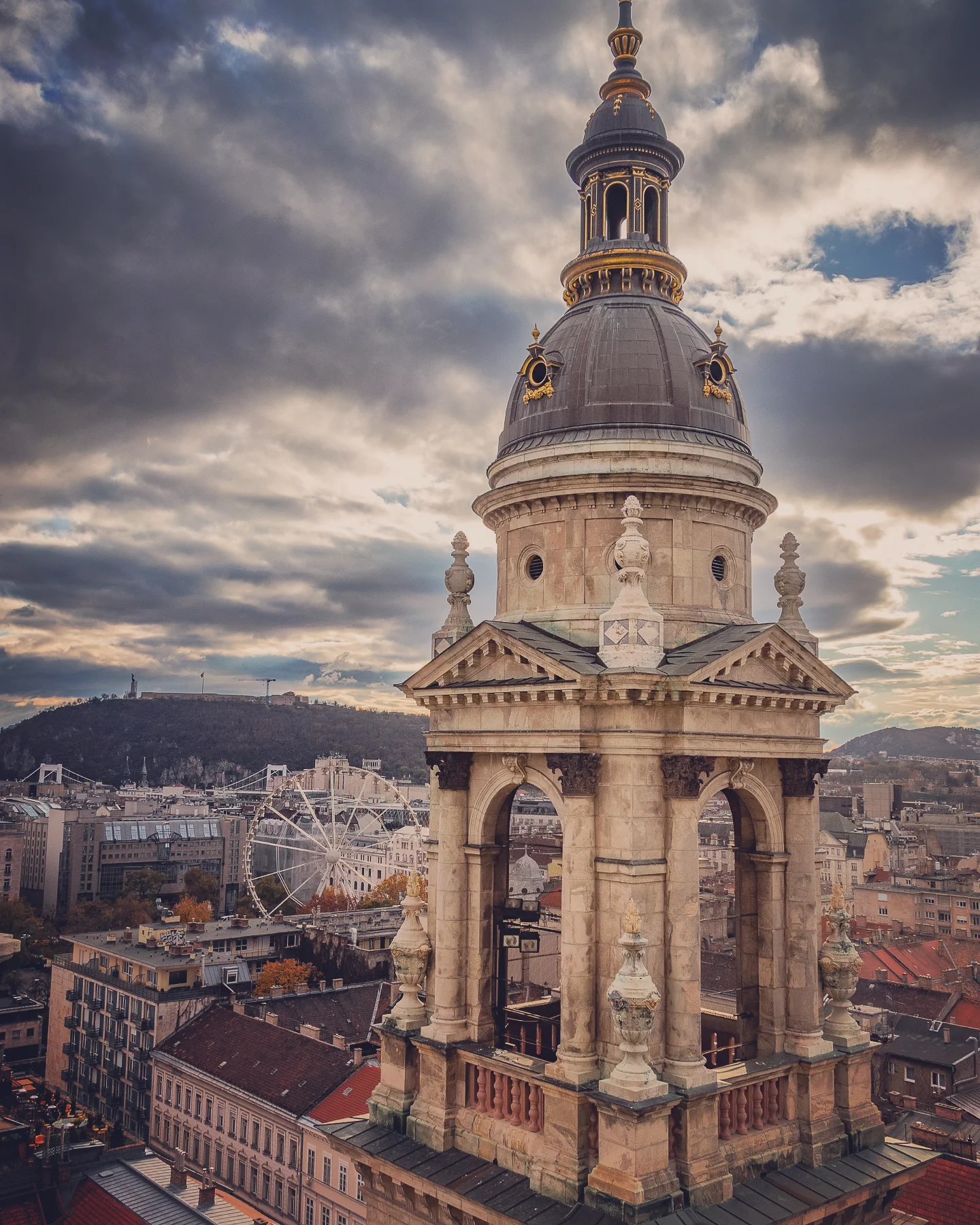 Dome and tower of St. Stephen’s Basilica in Budapest at sunset, with a Ferris wheel and cityscape in the background.