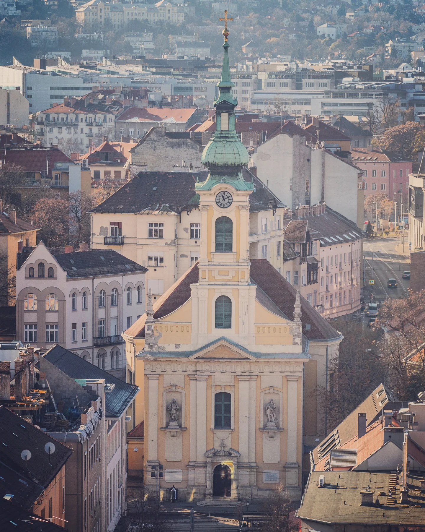 Yellow Baroque-style church with a green copper spire, surrounded by rooftops and city buildings.