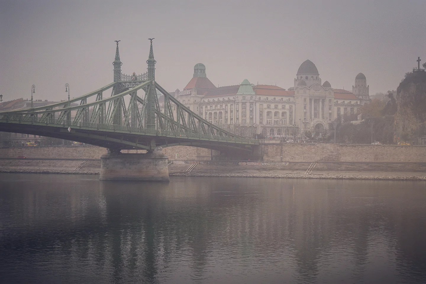 The green Liberty Bridge in Budapest spans the Danube River, with the grand Gellért Hotel and Baths rising behind it on a foggy morning.