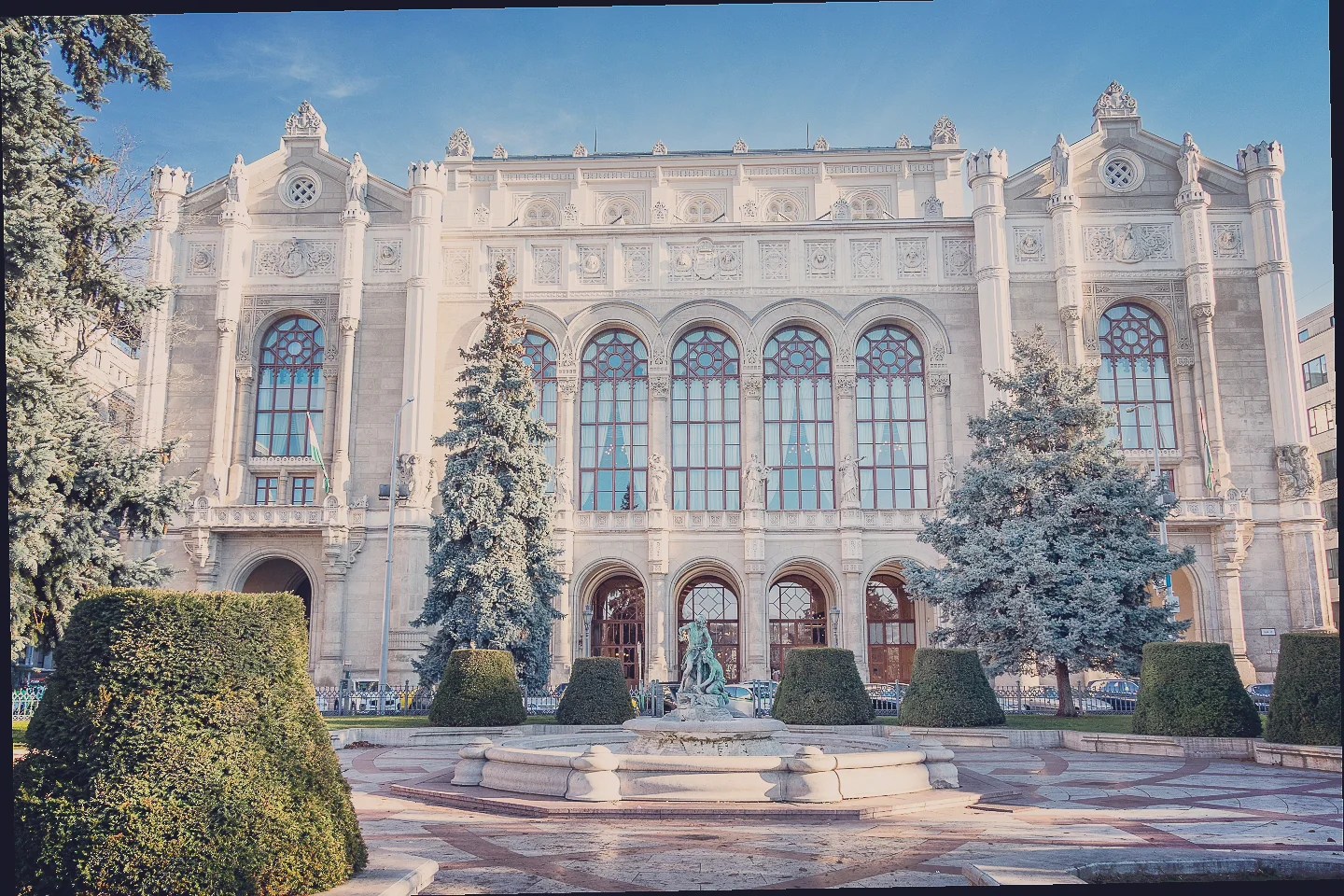 Ornate facade of Pesti Vigadó concert hall with large arched windows, statues, and a central fountain surrounded by manicured hedges.