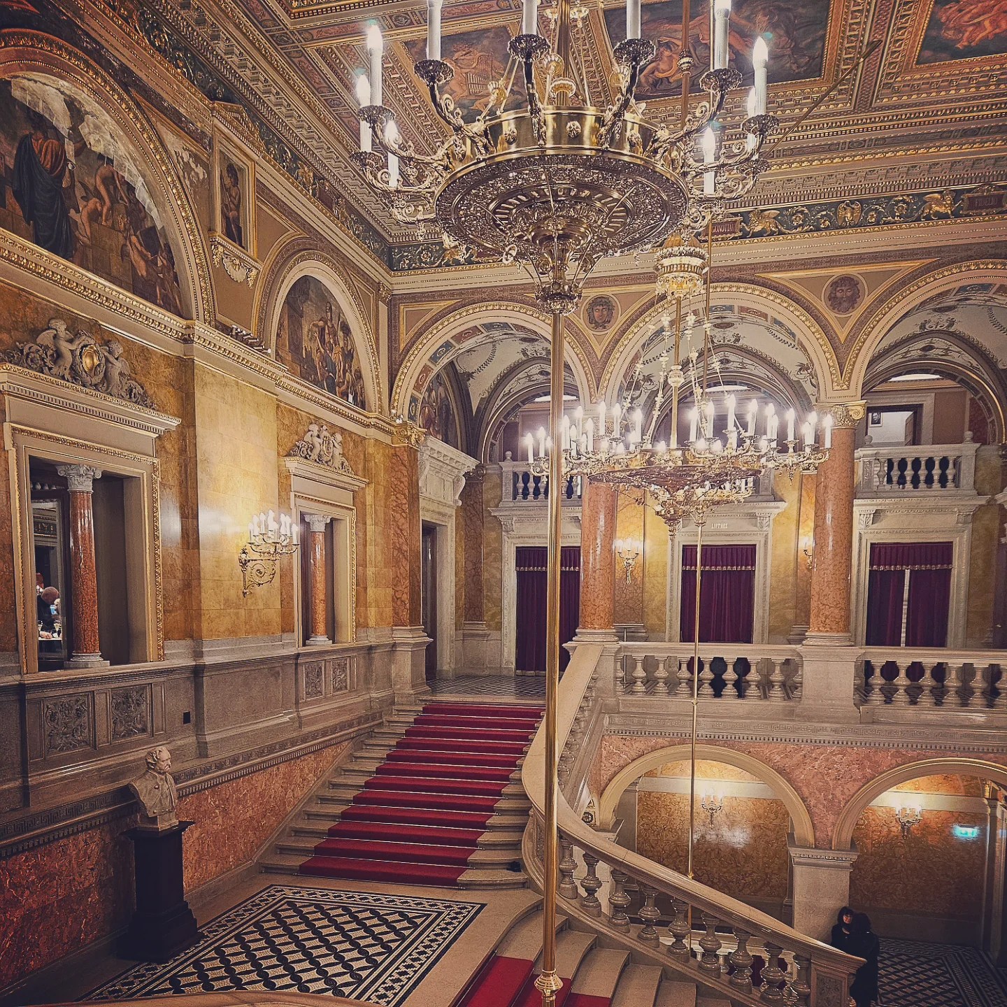 Ornate grand staircase inside the Hungarian State Opera House with chandeliers, red carpet, and painted ceilings.