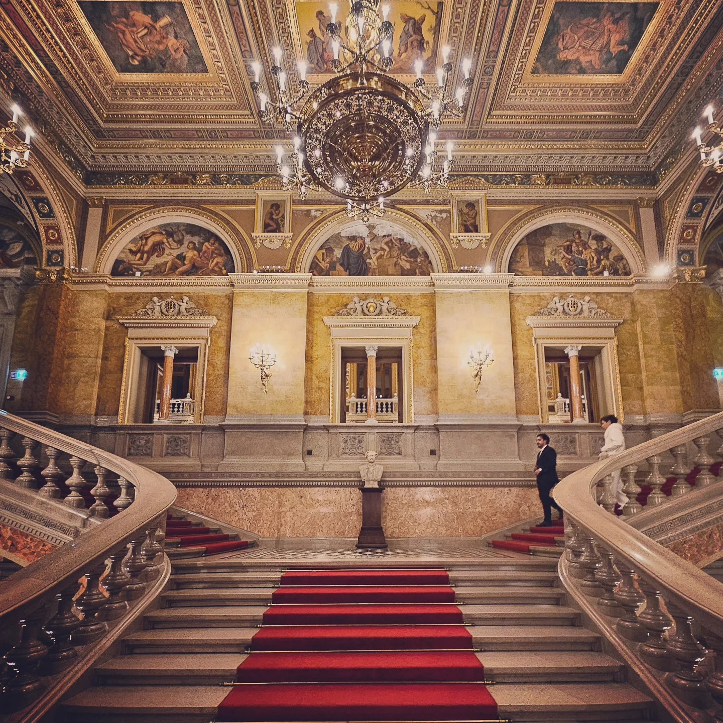 Central view of the grand staircase at the Hungarian State Opera House, framed by marble, murals, and a large chandelier.