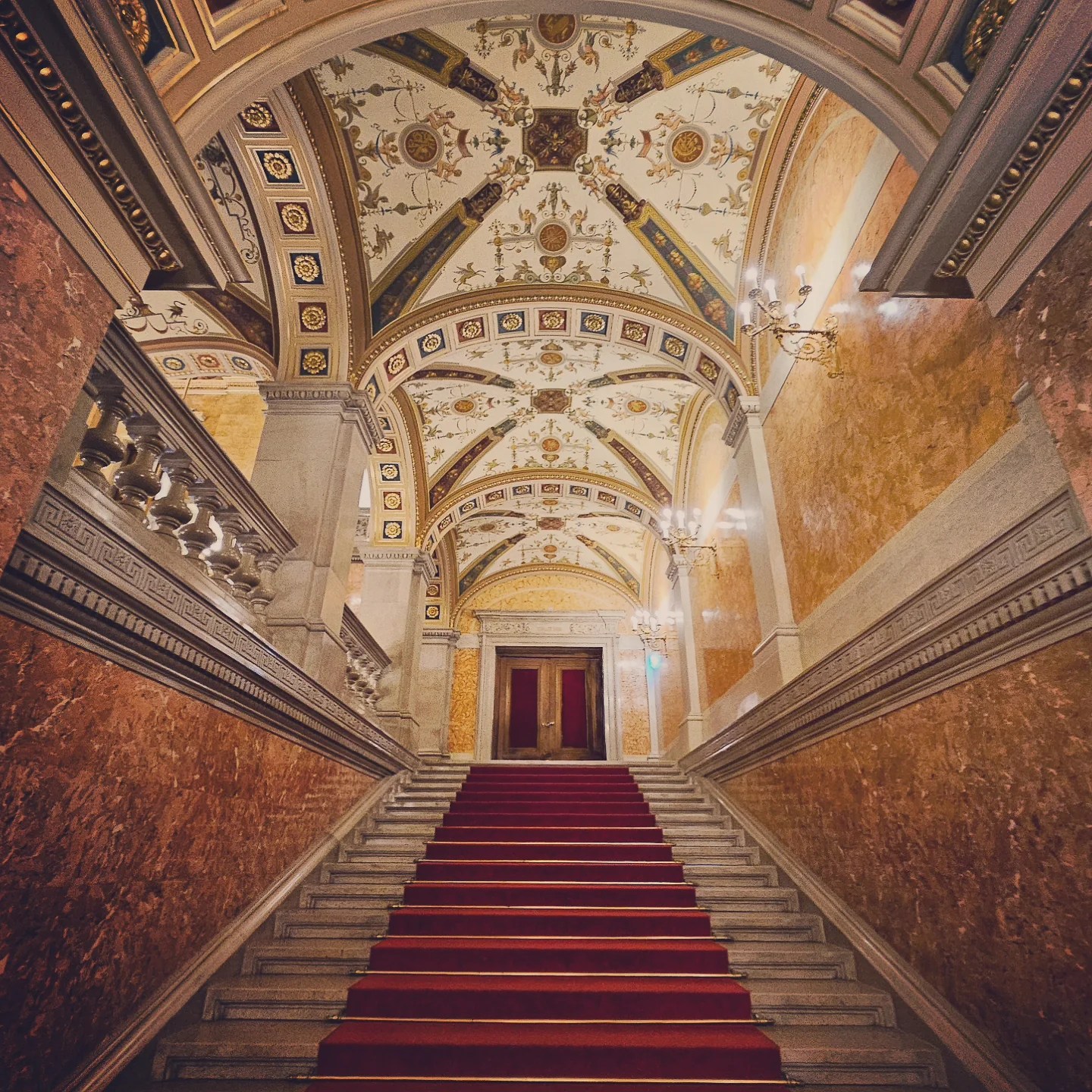 Ornate staircase with red carpet and a vaulted ceiling decorated with gold and floral details.
