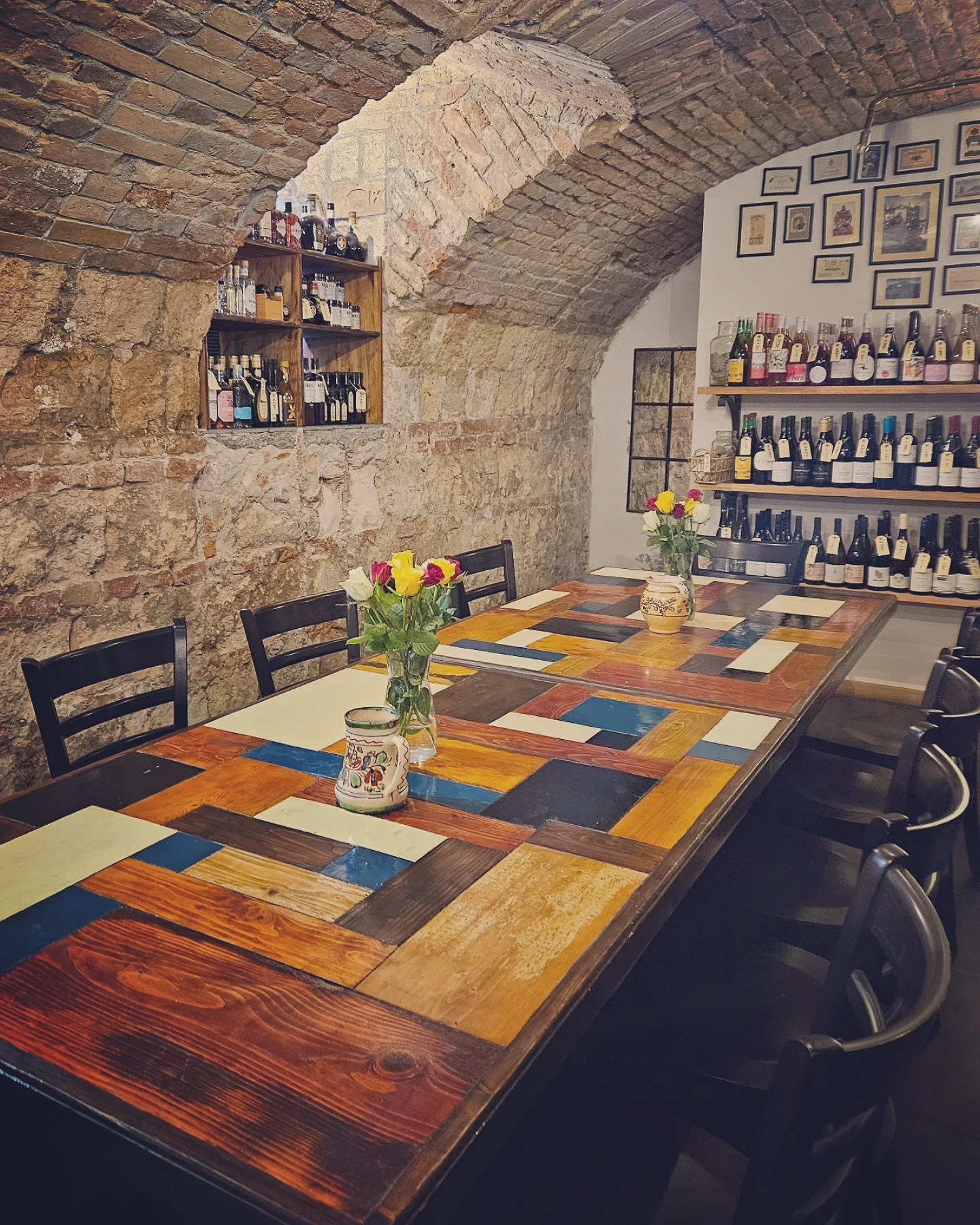 Long wooden table set with colorful tiles, surrounded by chairs and shelves of wine in a vaulted brick cellar.
