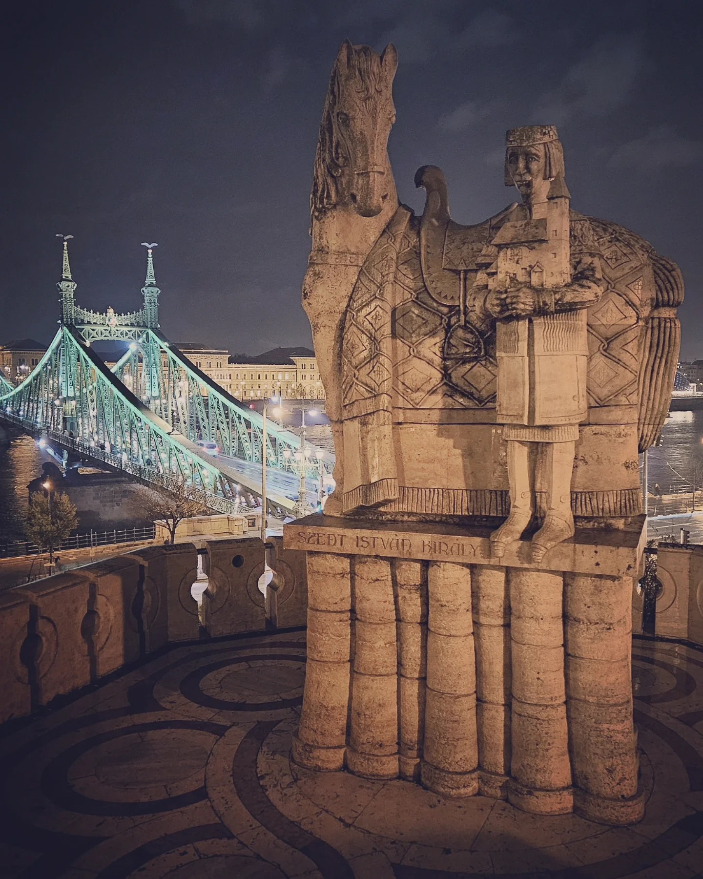 Stone statue of St. Stephen and his horse overlooking the illuminated Liberty Bridge in Budapest at night.
