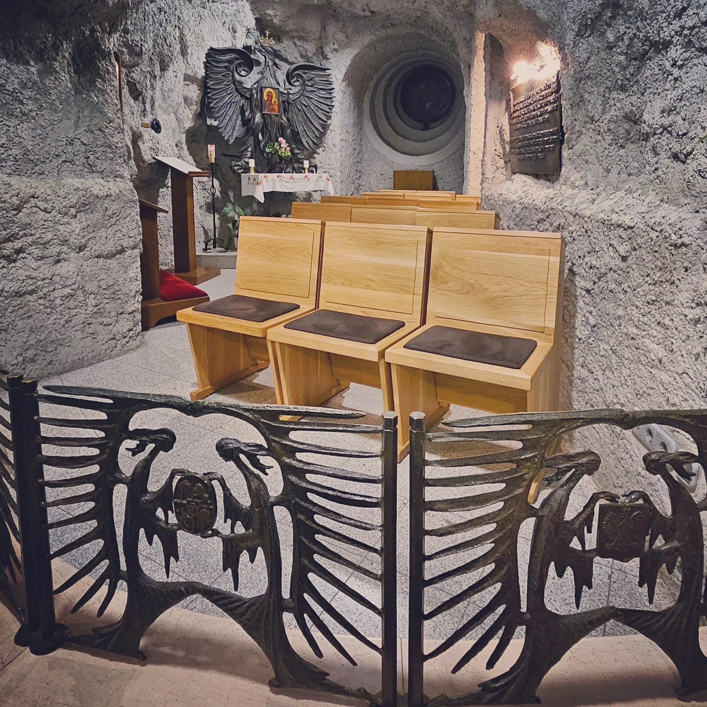 Interior view of Gellért Hill Cave Chapel in Budapest with natural stone walls, wooden pews, and an iron gate featuring winged women.