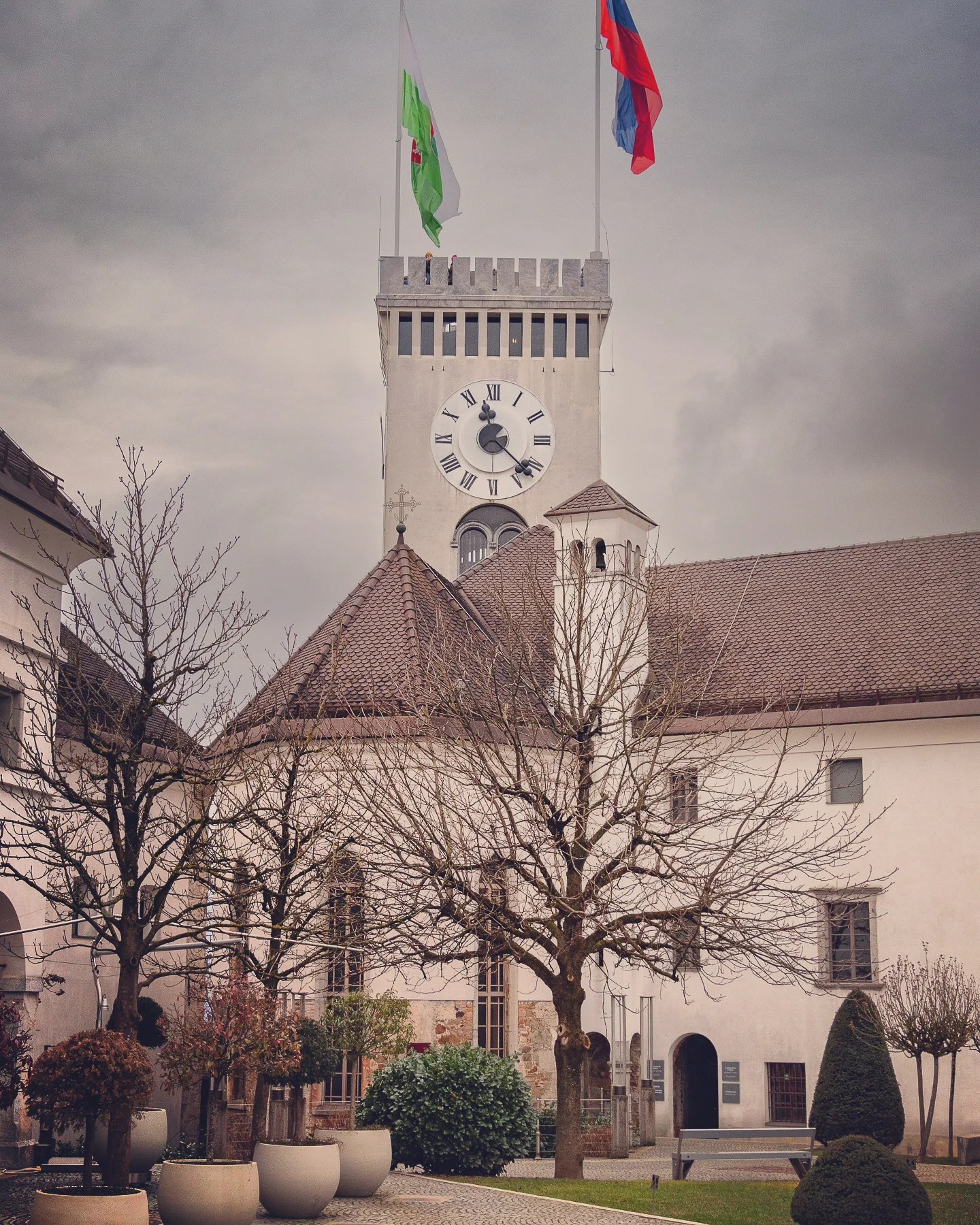 A view of Ljubljana Castle’s clock tower flying the Slovenian national flag and the city’s green dragon flag above a courtyard with leafless trees and potted plants under a cloudy sky.