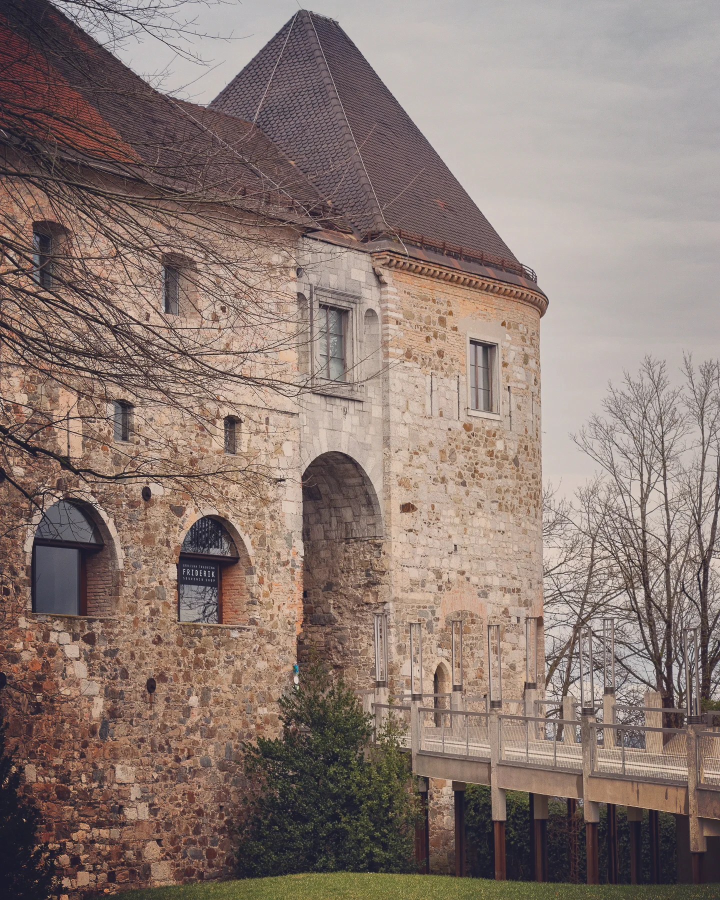 The entrance to Ljubljana Castle with a modern pedestrian bridge leading to a historic arched stone gate framed by bare winter trees.