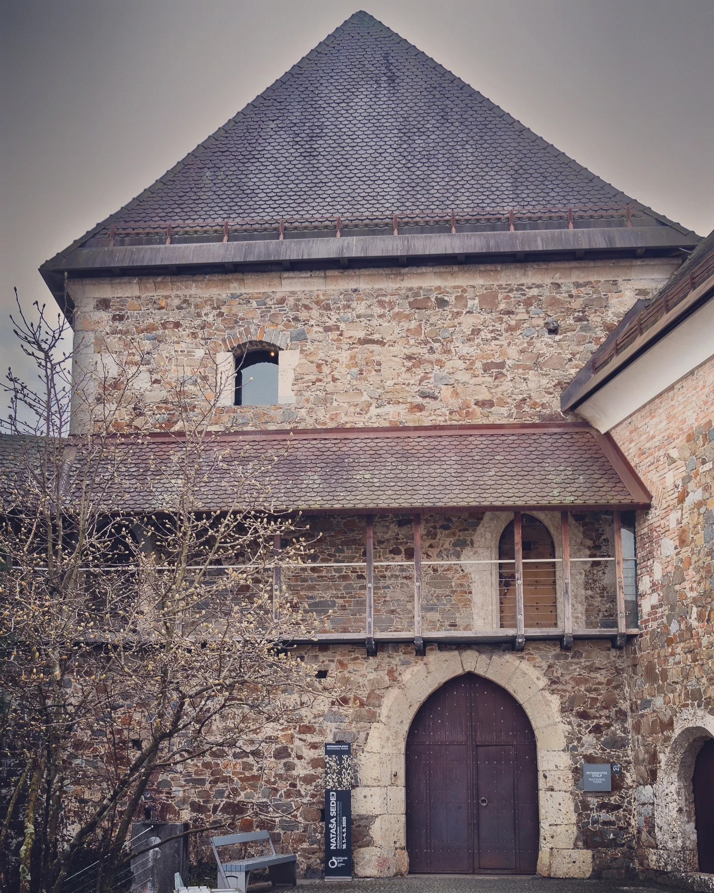 A fortified section of Ljubljana Castle with a pointed-roof tower, arched wooden door, and narrow balcony, surrounded by early spring trees.