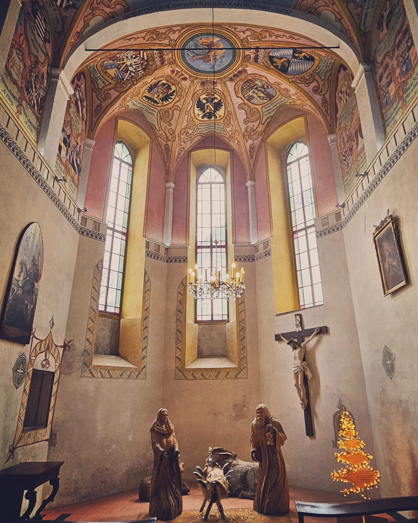 Interior of a richly decorated chapel at Ljubljana Castle, featuring colorful vaulted ceilings, stained glass windows, and a Christmas nativity scene with wooden figures.
