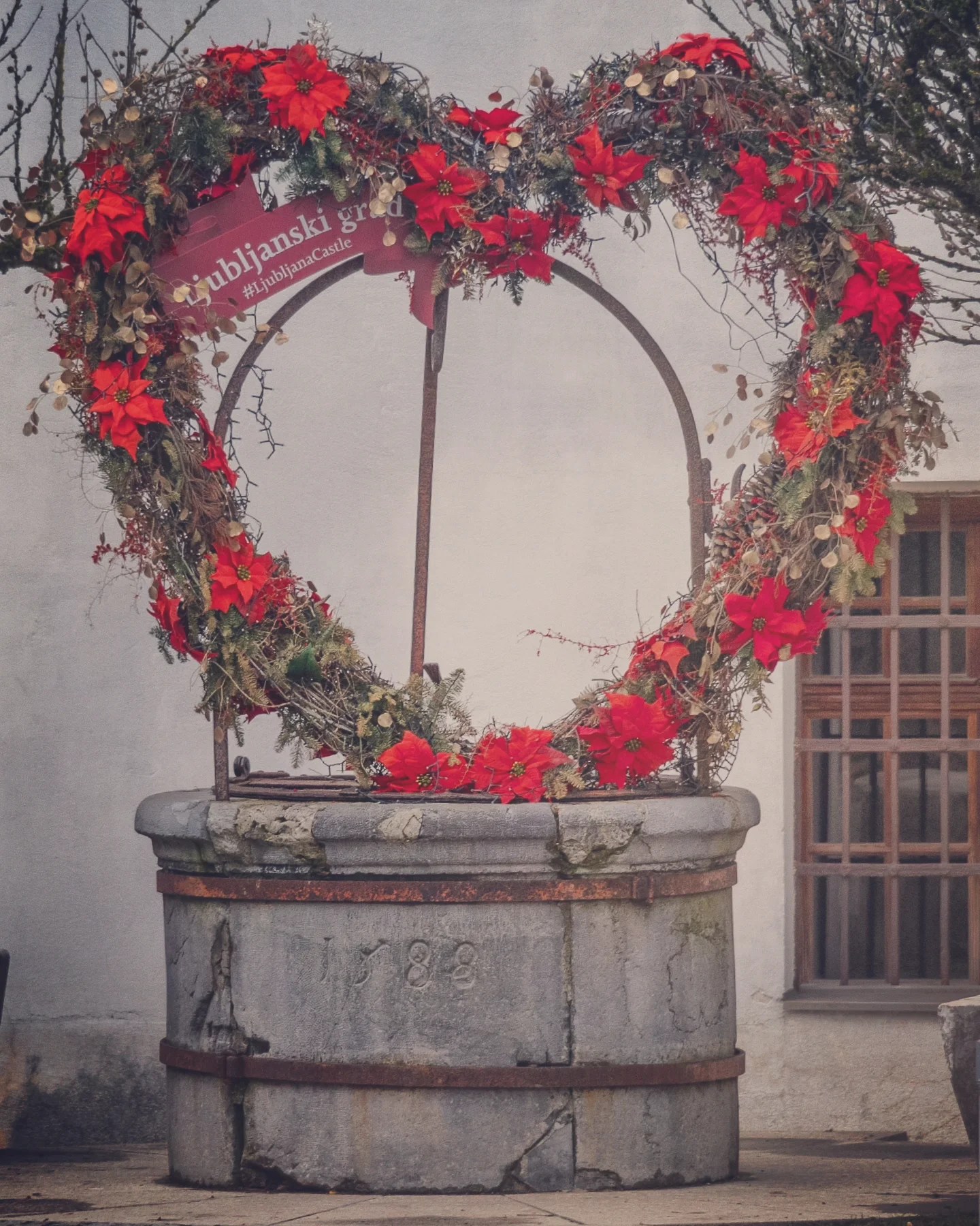 A festive heart-shaped floral wreath made of poinsettias and greenery framing a historic stone well dated 1588, marked with the sign “Ljubljanski grad” and hashtag “#LjubljanaCastle.”