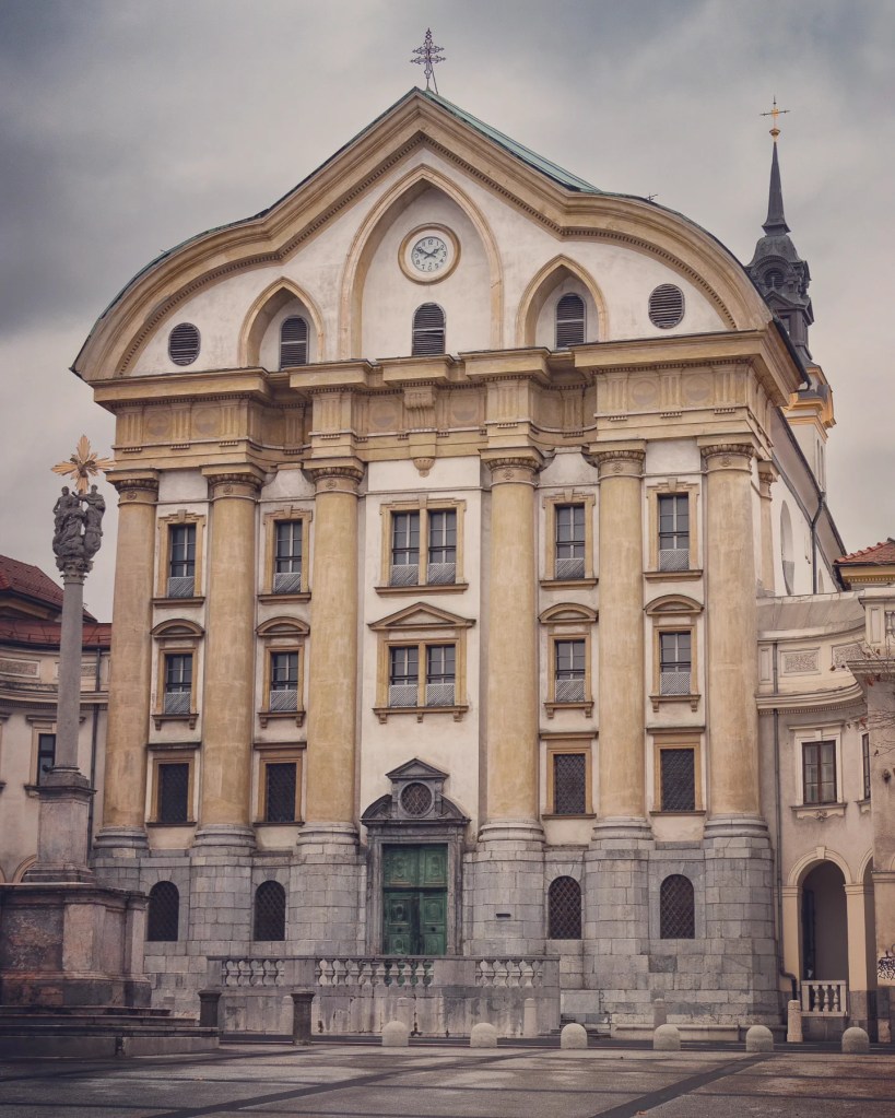 The elegant Baroque façade of the Ursuline Church of the Holy Trinity in Ljubljana, featuring a pale yellow and white exterior with columns, a clock, and a cross-topped gable under a cloudy sky.