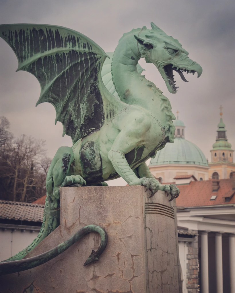A side view of Ljubljana’s famous green dragon statue on Dragon Bridge, snarling with wings raised and a backdrop of domed rooftops and historic architecture.