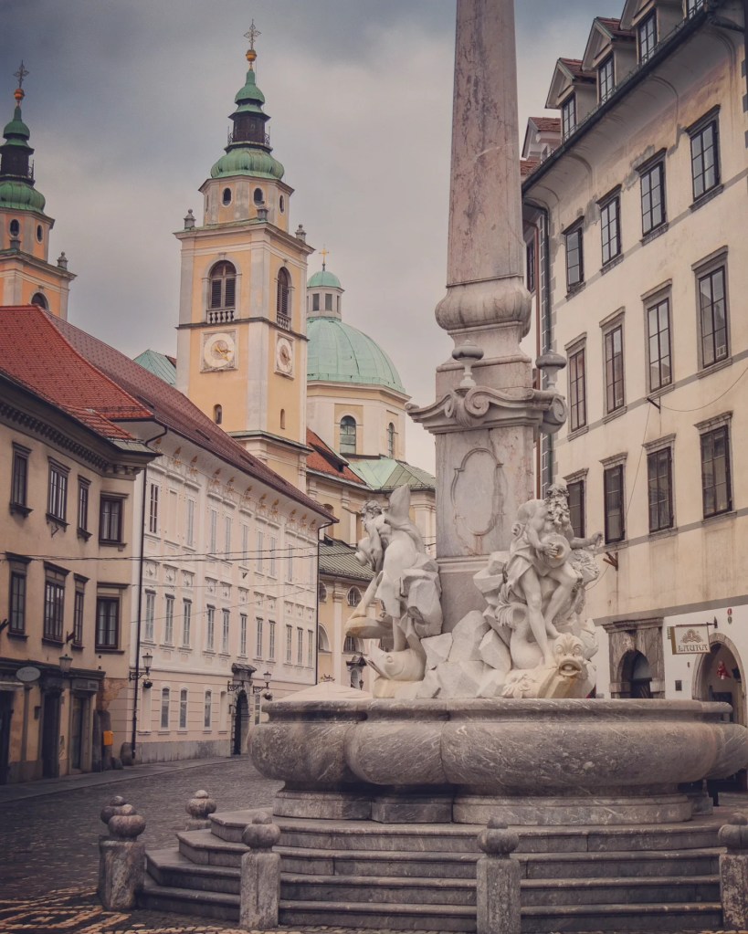 The Robba Fountain in Town Square, Ljubljana, adorned with marble sculptures of mythological figures, set against the backdrop of the Ljubljana Cathedral’s twin green-domed towers.