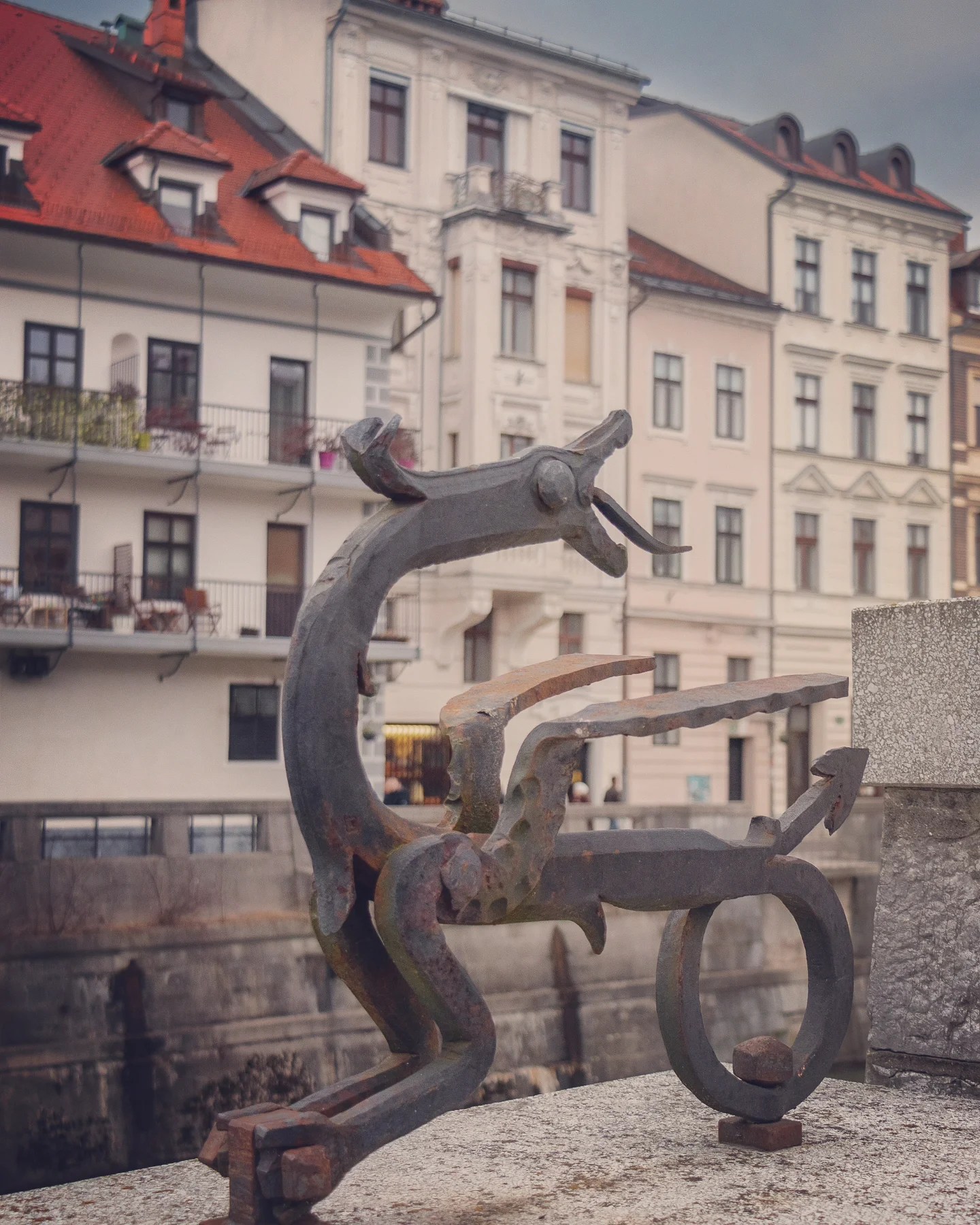 A whimsical iron dragon sculpture perched on the edge of a wall, with pastel-colored buildings and red rooftops in the background along the Ljubljanica River.