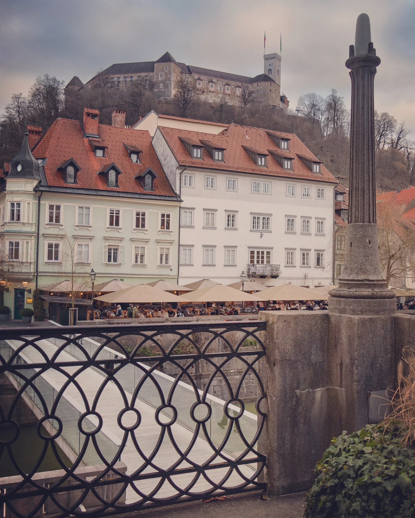 A scenic view of Ljubljana Castle on the hilltop above the old town, with red-roofed houses and bustling riverside cafés in the foreground, seen through decorative iron railing.