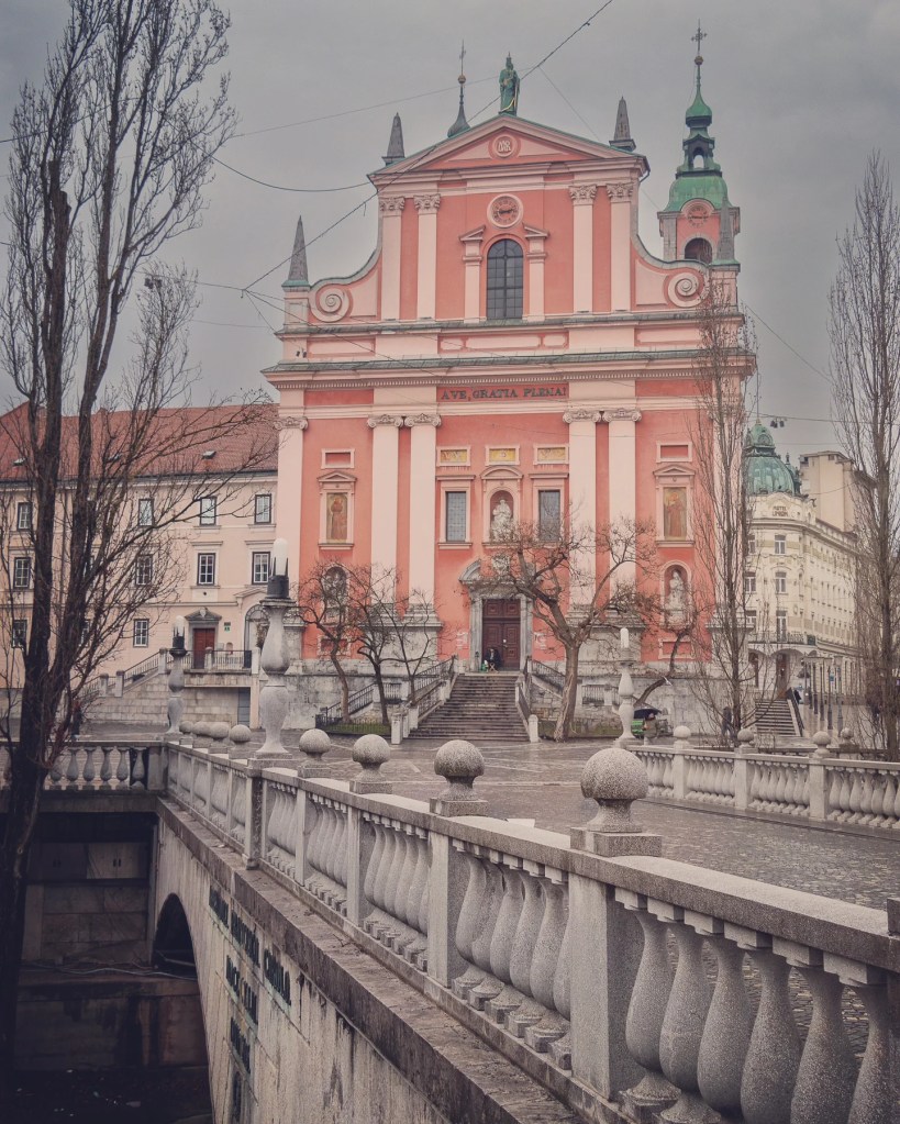 A wider view of Ljubljana’s iconic Franciscan Church with its coral pink façade, seen from across the Triple Bridge with decorative stone balustrades leading toward the entrance.