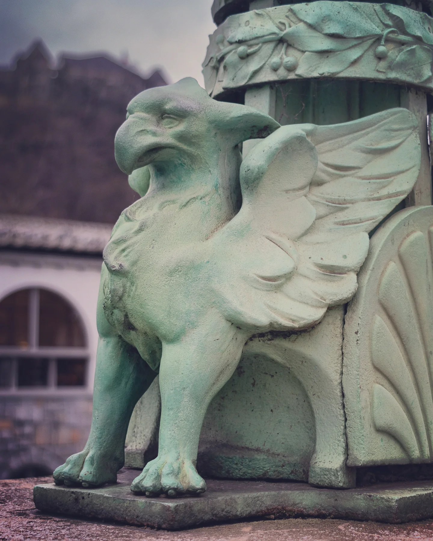 A close-up of a small green griffin statue with feathered wings and a strong beak, perched on a decorative pedestal near the Dragon Bridge in Ljubljana.