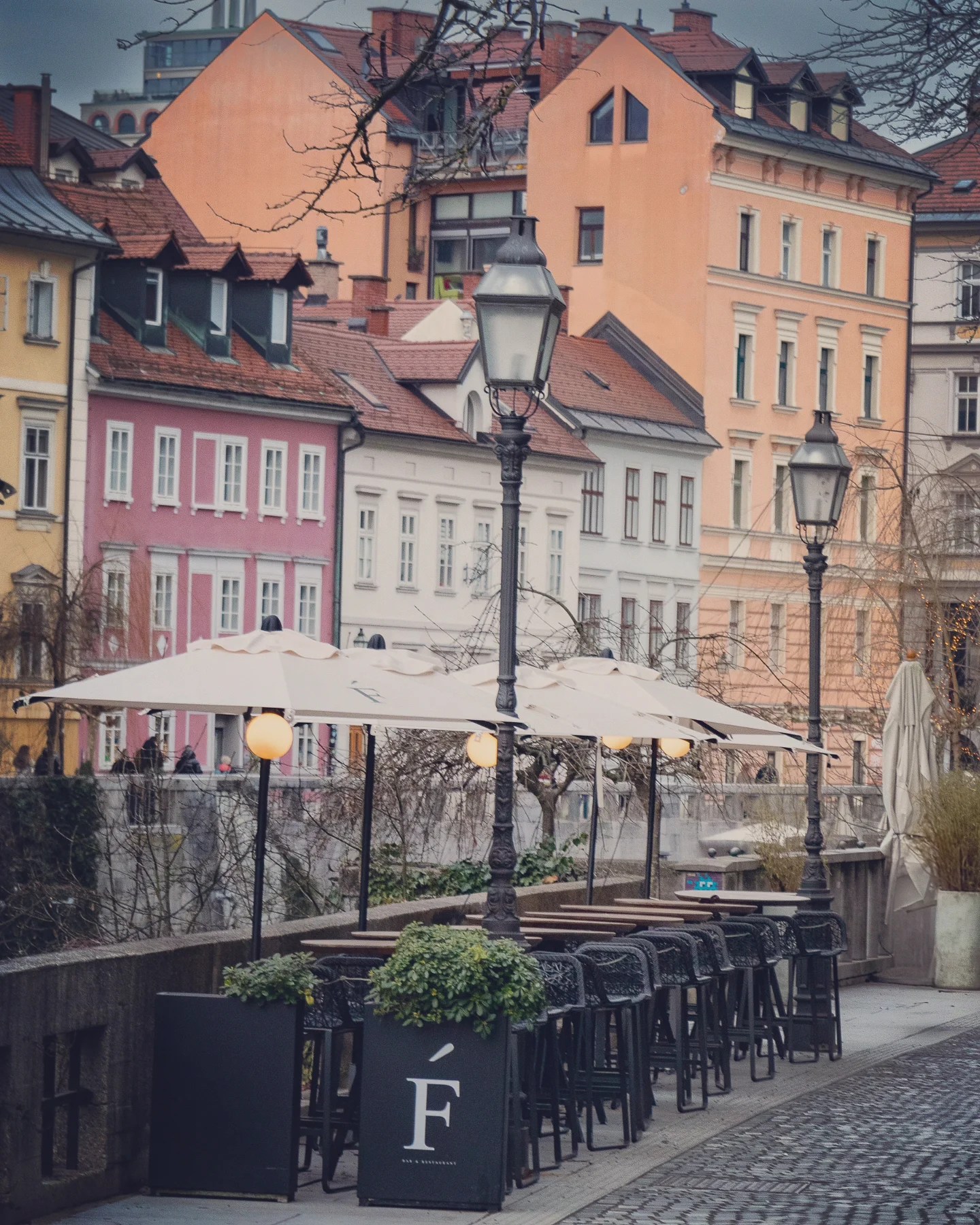 Outdoor seating at a stylish café along the Ljubljanica River, lined with white umbrellas and vintage streetlamps, backed by a vibrant row of pastel-colored buildings.