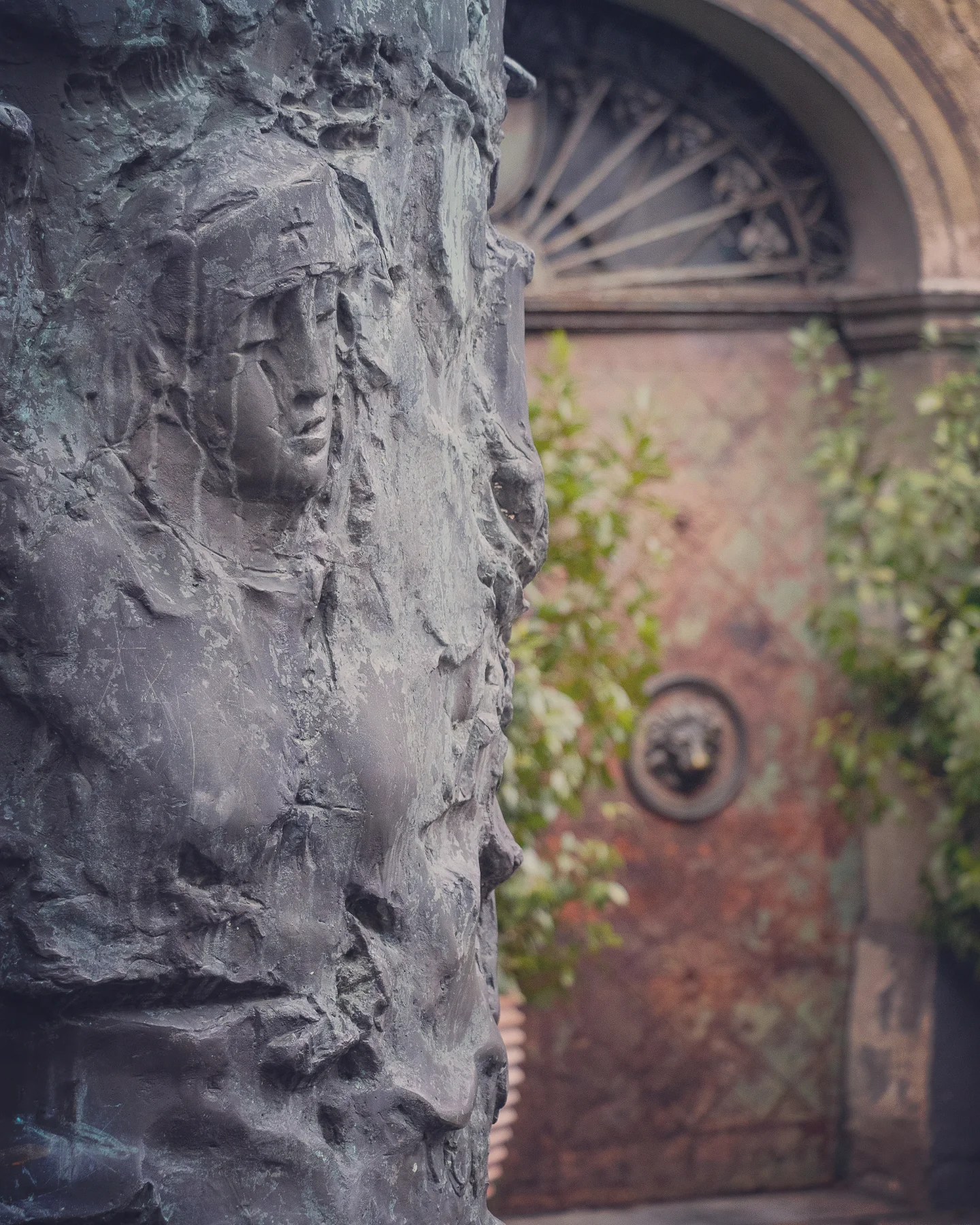 A textured bronze column featuring expressive human faces and figures emerging from the surface, located in front of a historic doorway with ornate ironwork.
