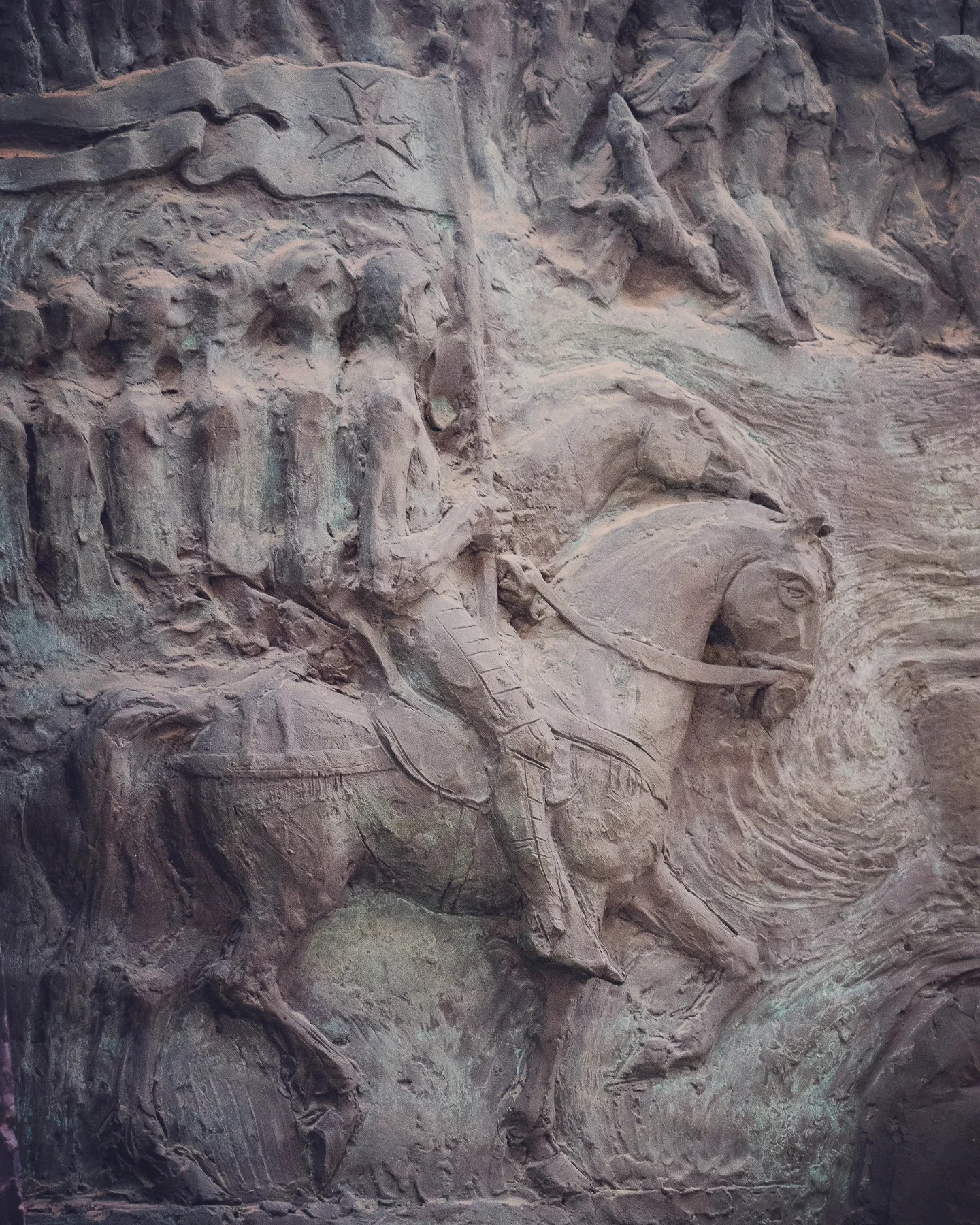 A close-up of a dramatic bronze bas-relief sculpture depicting a mounted figure holding a flag, surrounded by a procession of soldiers and dynamic movement.
