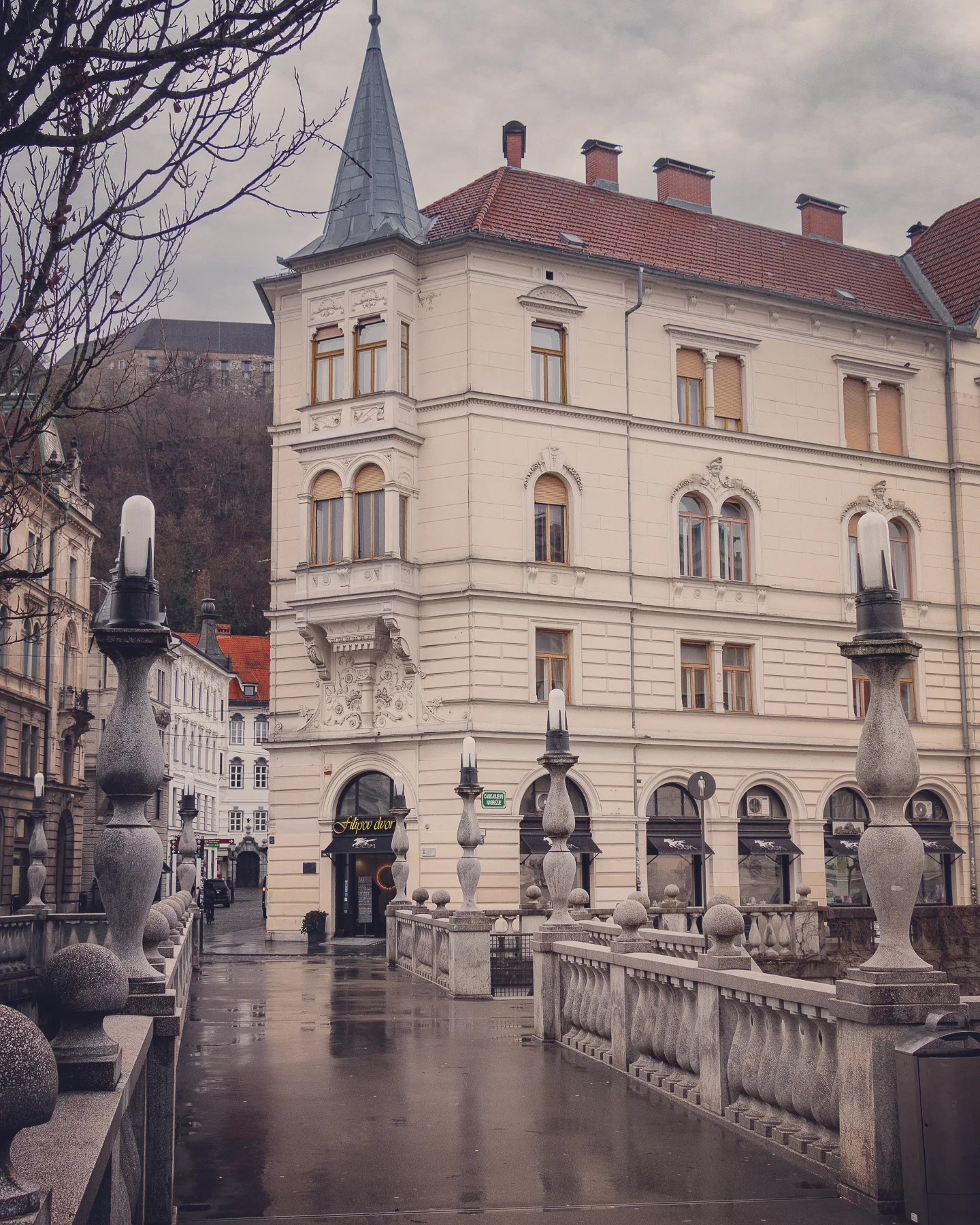 A view of a cream-colored historic building with arched windows and a pointed turret, framed by stone balustrades and lamps along the Triple Bridge on a rainy day.