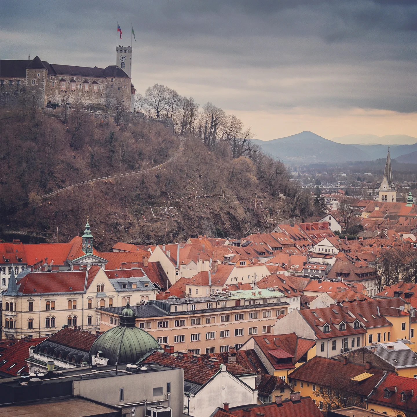 A panoramic view of Ljubljana Castle atop a forested hill, overlooking the terracotta rooftops and domes of the old town under a moody winter sky.
