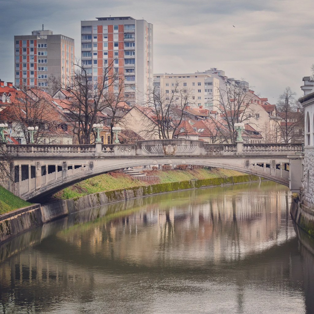A graceful stone bridge engraved with "1848" and "1888" crossing the Ljubljanica River, surrounded by mid-century apartment buildings and reflecting in the water below.