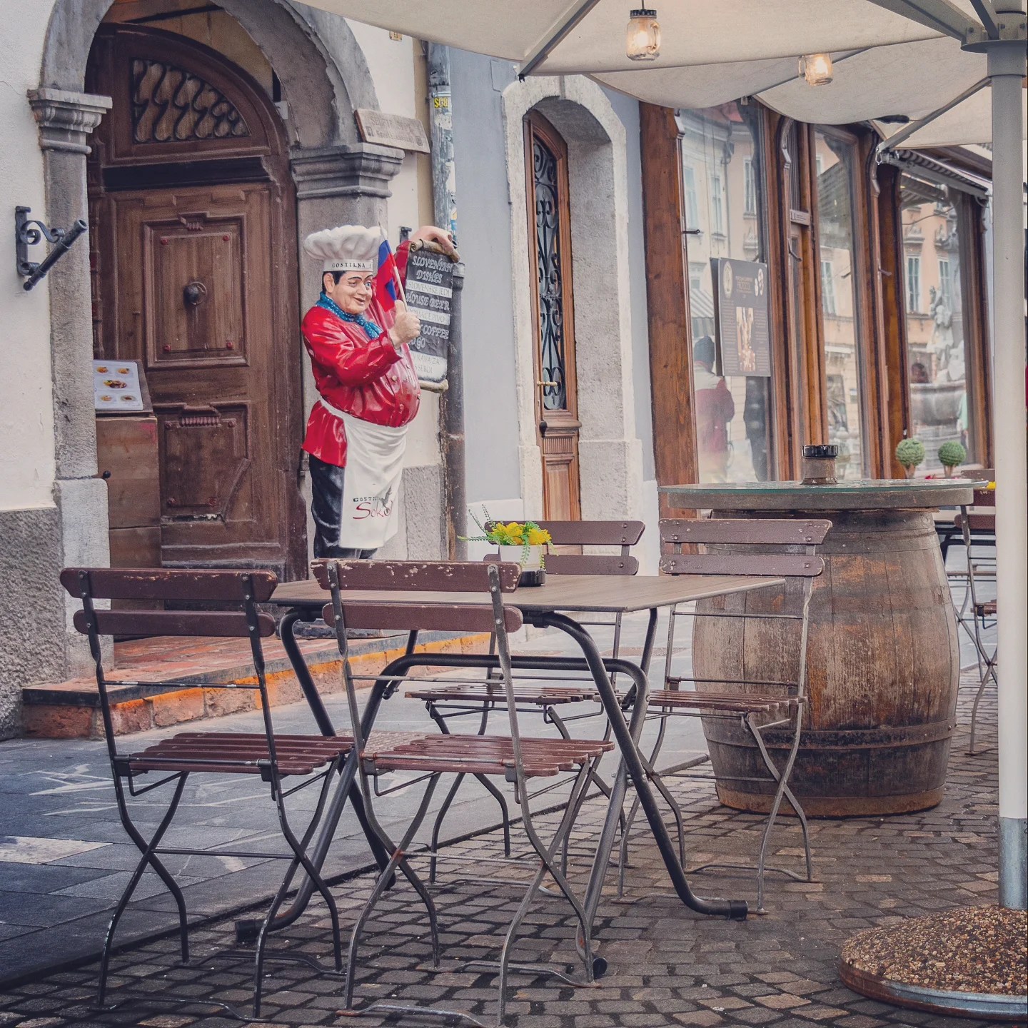 A quiet café terrace with wooden tables and chairs beneath white umbrellas, watched over by a whimsical life-sized statue of a smiling chef holding a menu board.