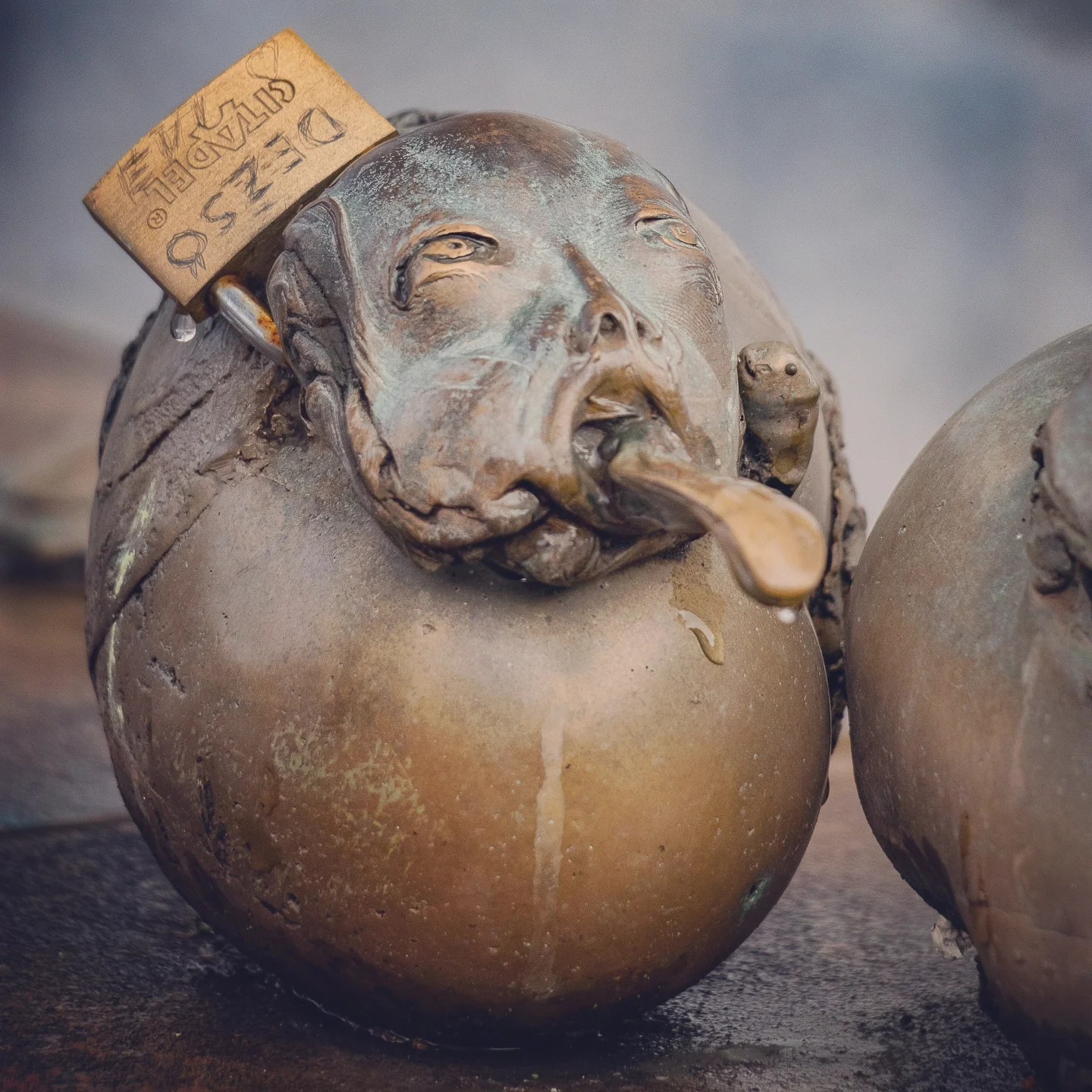 A close-up of a bizarre bronze head sculpture with a tongue sticking out, next to a weathered lock engraved with a message—an example of Ljubljana’s quirky public art.