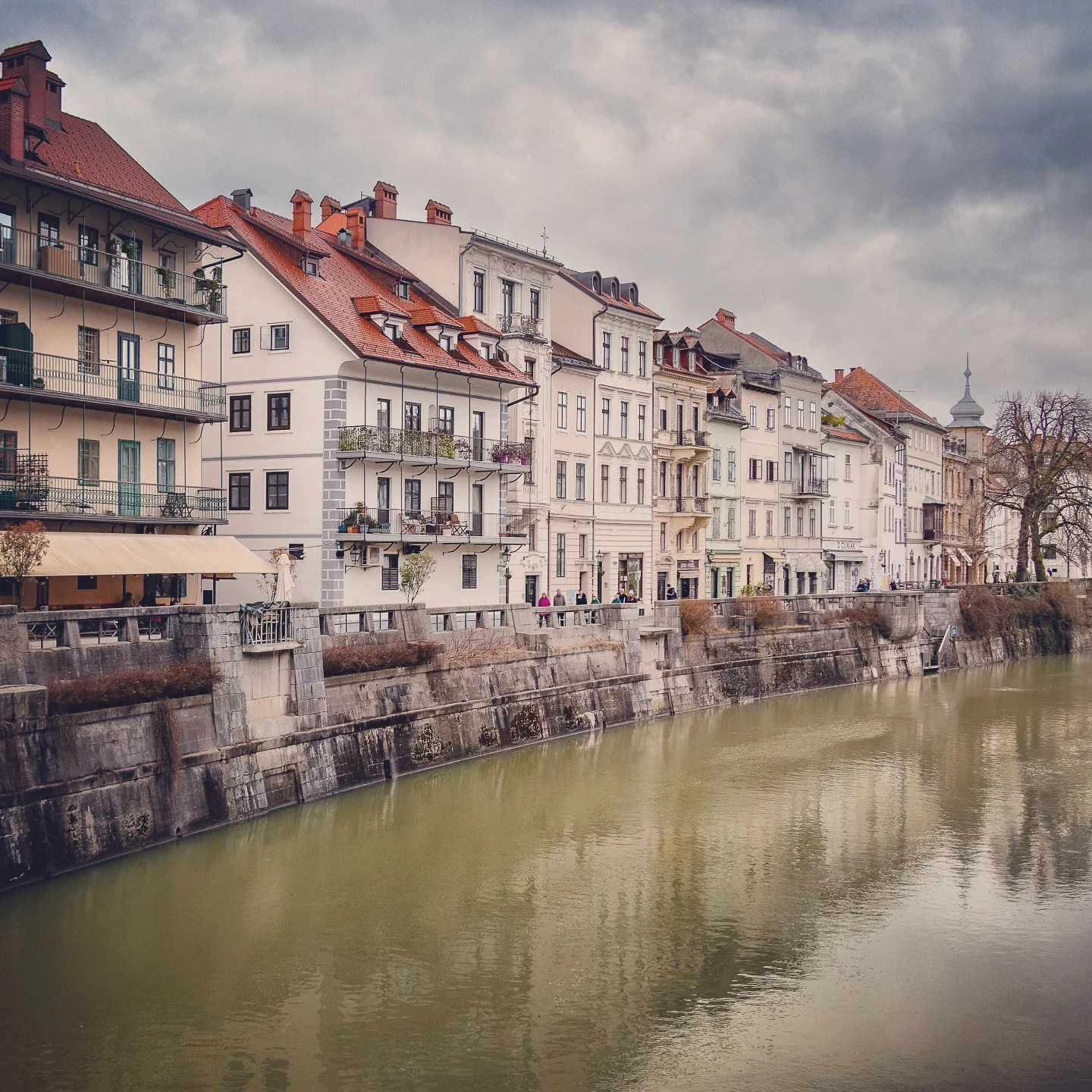 A moody riverside view of cream and pastel-colored buildings with classic European architecture lining the Ljubljanica River under a cloudy winter sky.