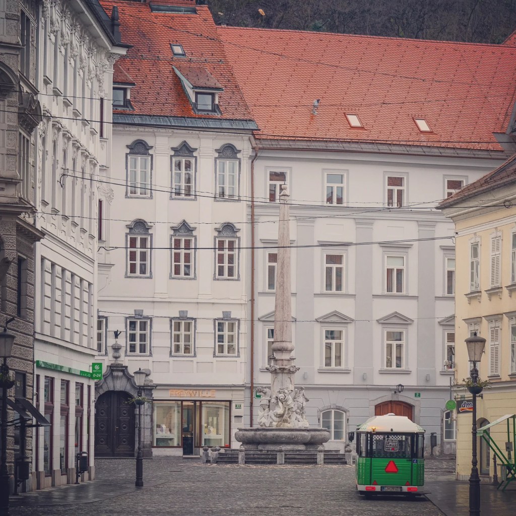 Ljubljana’s free green electric tourist train parked near Robba Fountain in a quiet cobblestone square surrounded by white and yellow historical buildings.