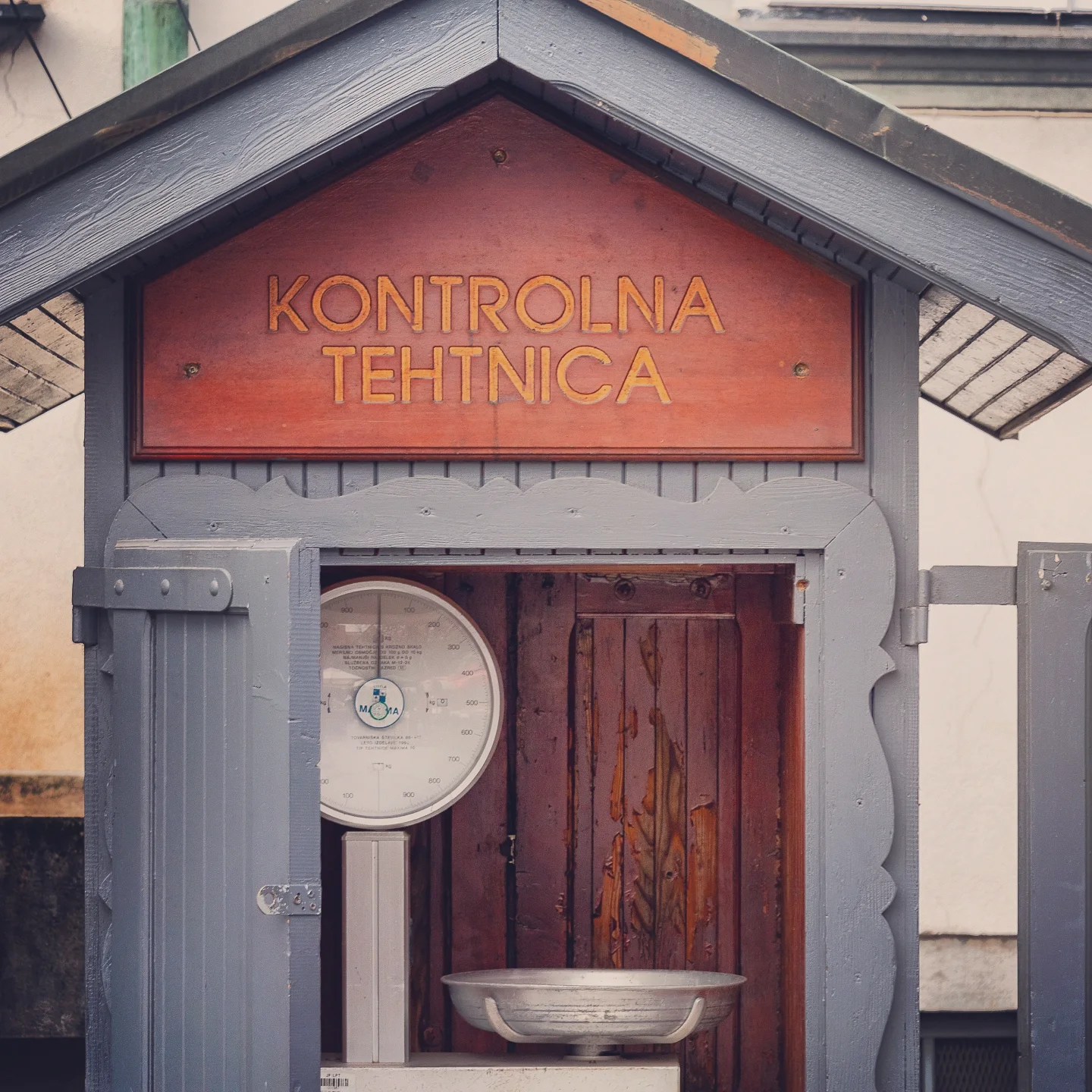 A small wooden hut painted blue and red housing a vintage public weighing scale, marked “Kontrolna Tehtnica.”
