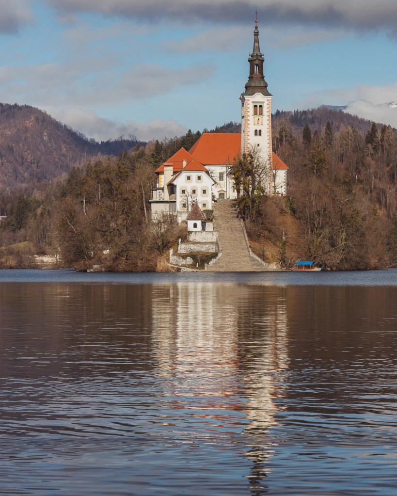 The Church of the Mother of God on Bled Island reflected in the still waters of Lake Bled, framed by wooded hills under a partly cloudy sky.