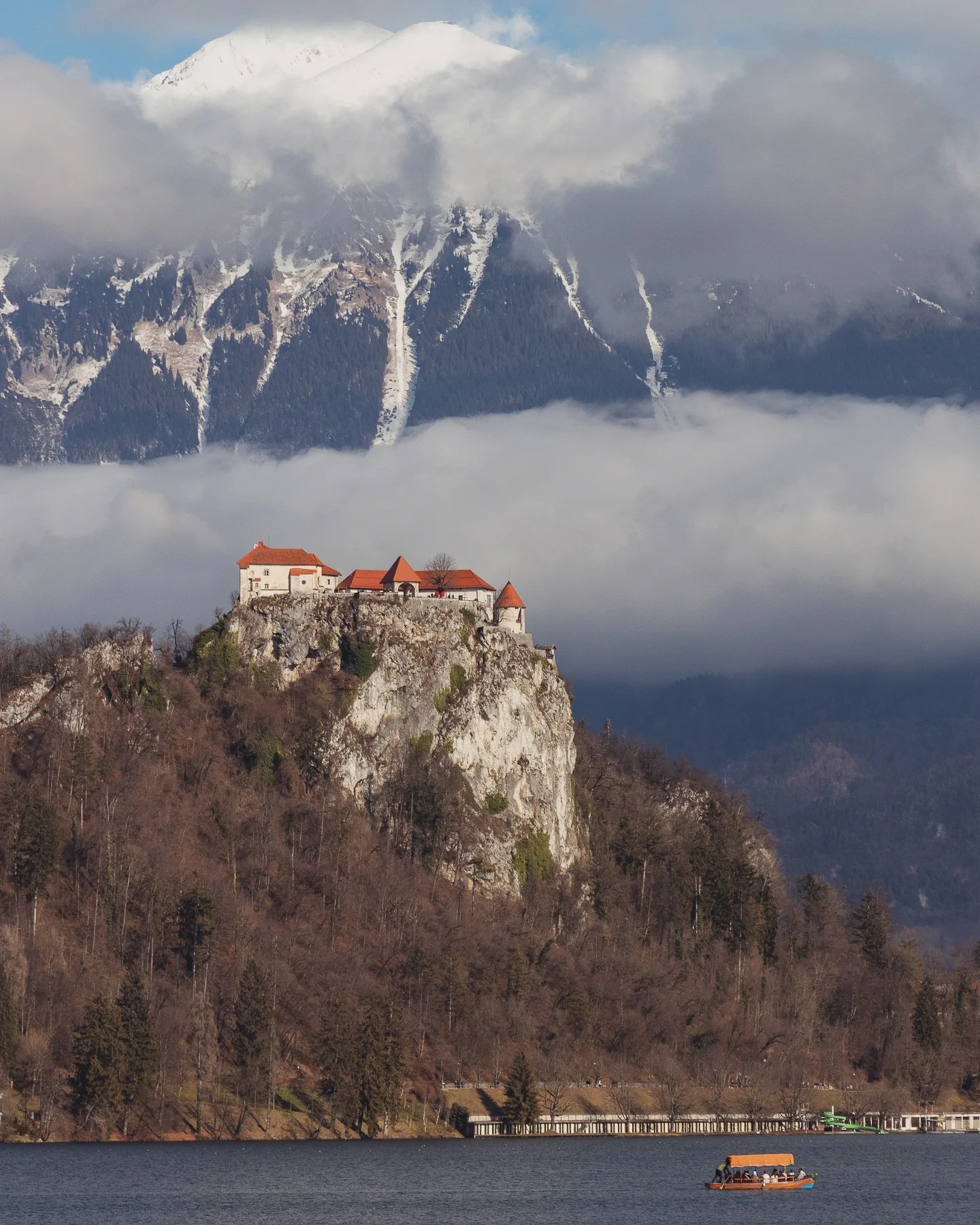 Bled Castle rising dramatically from a steep cliff above Lake Bled, with a traditional wooden boat floating on the lake and snowy peaks looming in the background.