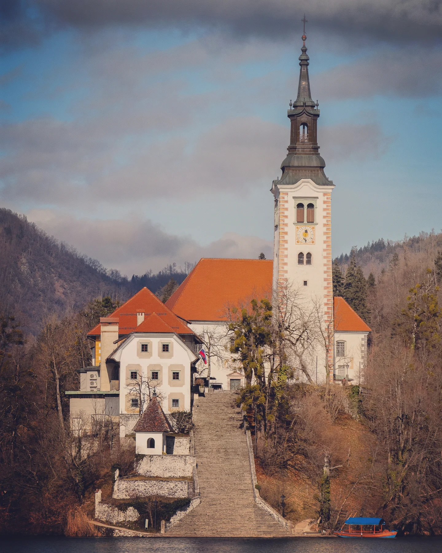 Close-up view of the Church of the Assumption on Bled Island, showing the steep staircase leading to the entrance and the church tower rising into a partly cloudy sky.