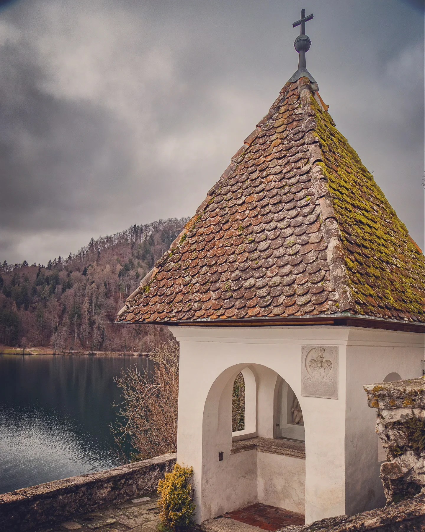 A small white chapel with a mossy tiled roof and a cross on top, overlooking the tranquil waters and wooded hills of Lake Bled on a cloudy day.