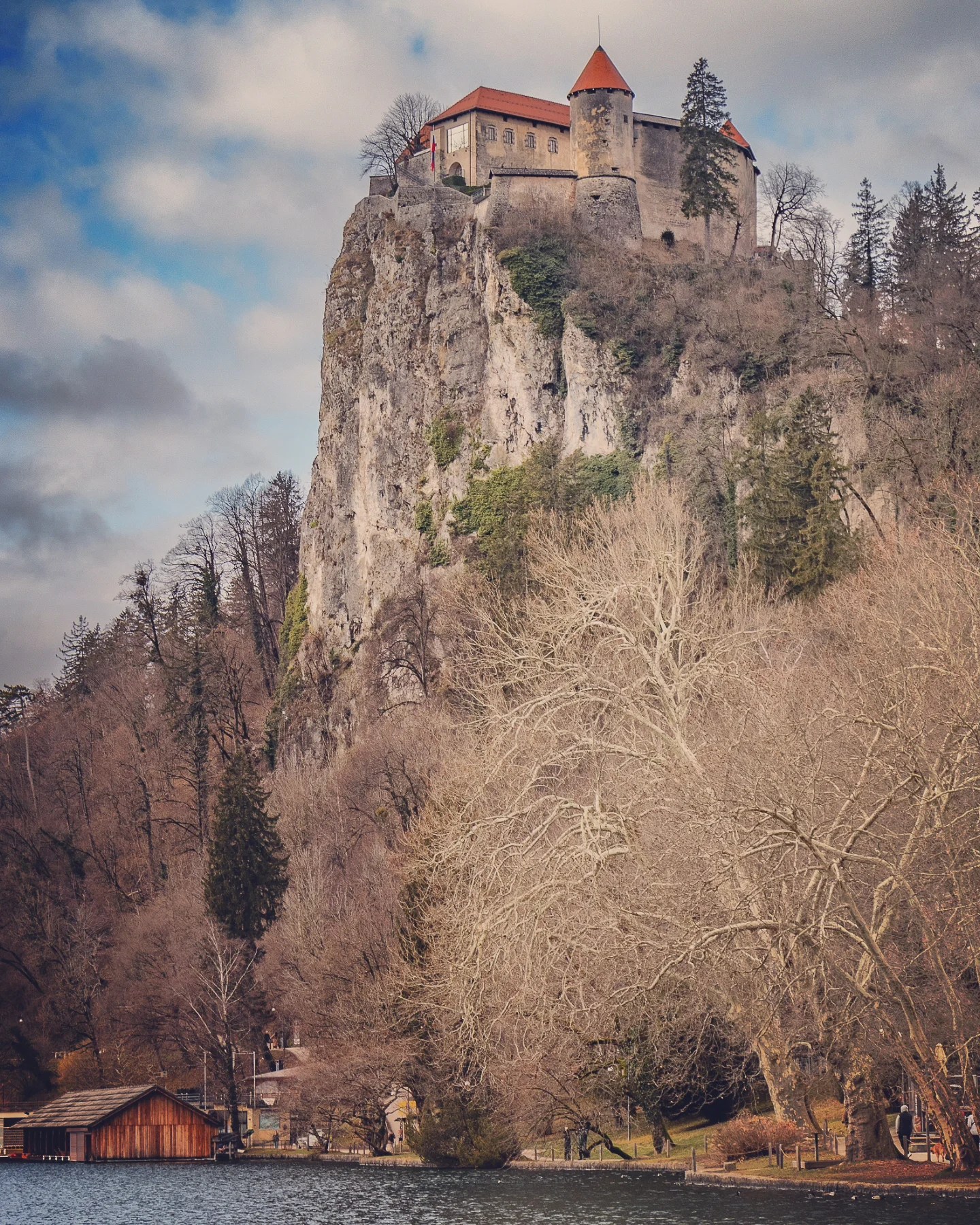 A close-up perspective of Bled Castle clinging to a rocky cliff above the lake, surrounded by leafless trees and framed by a partly cloudy blue sky.