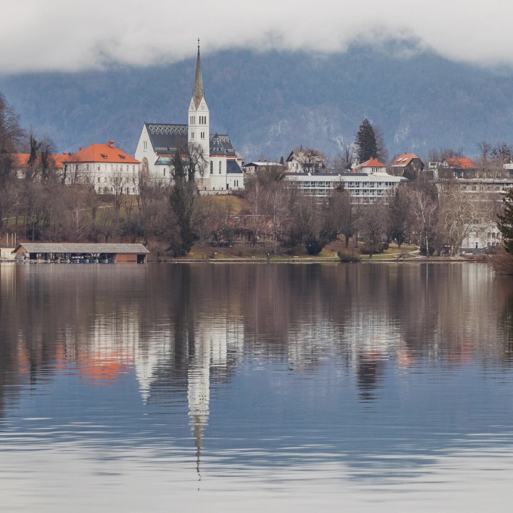 Reflections of a white church with a tall spire and red-roofed buildings on the calm surface of Lake Bled, backed by misty forested mountains.