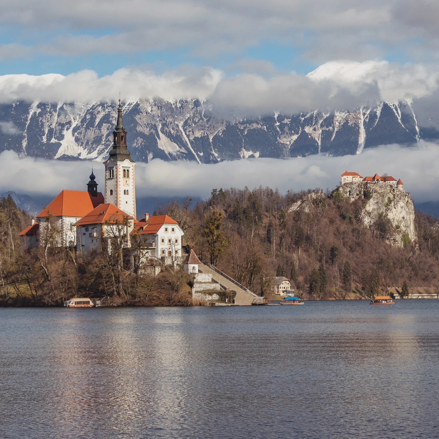 Iconic view of Lake Bled with the Pilgrimage Church of the Assumption on Bled Island in the foreground and Bled Castle perched on a cliff in the distance, all set against snow-capped Julian Alps.
